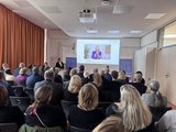 Veranstaltungsraum Publikum von hinten. Blick auf eine Beamer-Leinwand, auf der die Bayerische Staatsministerin für Unterricht und Kultus, Anna Stolz, zu sehen ist.