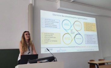 Symposium auf der PAEPS Antonia Landgraf an einem Rednerpult mit großer Beamer-Präsentation im Hintergrund